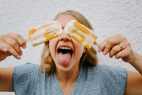 Female making funny grimace with tongue out and covering eyes with tasty ice lollies on sticks