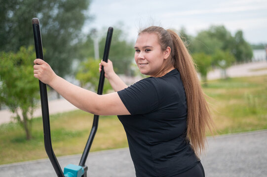 Beautiful smiling overweight young woman does fitness on an ellipsoid simulator outdoors. Fat girl is training on the sports ground for weight loss. Sport outside on a warm summer day.