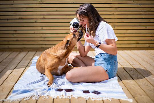 Side view of positive young female in casual shirt and jeans shorts taking pictures with instant camera of cute dog lying nearby white resting on sunny terrace in summer day