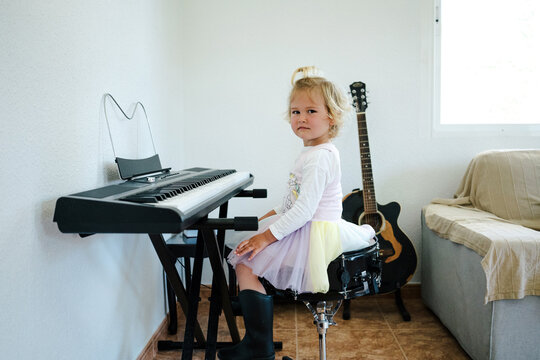 Side view of schoolgirl in fluffy skirt sitting at synthesizer and preparing for music class looking at camera