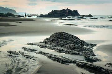 Sea waves rolling on wet sandy seashore with rough rocky formations and distant silhouette of female traveler in summer evening in Taiwan