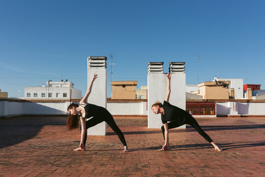 Full body side view of slim women in black activewear performing Revolved Triangle Pose while practicing yoga together on rooftop terrace in city