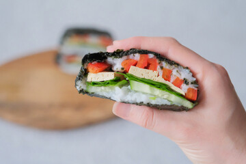 The hand of young woman, holding  onigirazu, vegan sushi sandwich with vegetables (cucumber, pepper, spinach) and tofu. Trend Japanese food. Healthy food concept.