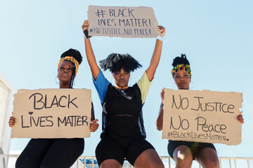Group of multiethnic female activists protesting with raised arms in city with carton placards with black lives matter inscription and looking at camera