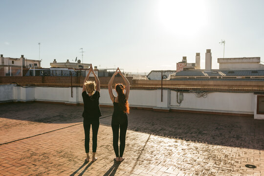 Full body back view of mature woman with young daughter in black sportswear standing in Upward Salute position with raised arms while doing Sun Salutation sequence during yoga practice on rooftop