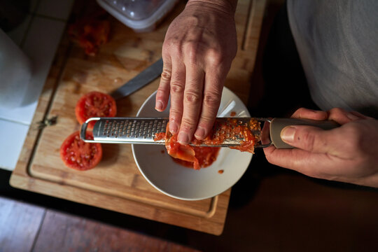 Stock photo of an aerial view of hands of a man grating tomato on top of a piece of wood in a kitchen