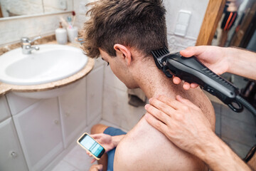 From above side view of crop shirtless male focusing on screen and watching tutorial video after self cutting hair in light bathroom at home as concept bad decisions or result of self isolation