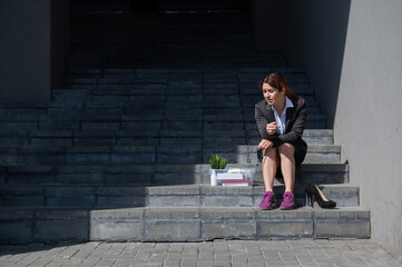 Depressed woman dressed in medical mask is fired and is sitting on the stairs with a box of personal belongings. Female office worker in suit and sneakers outdoors. Unemployment in the economic crisis