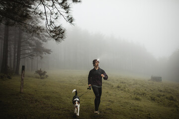 Confident male runner in sportswear training in misty wood with dog while jogging and looking away