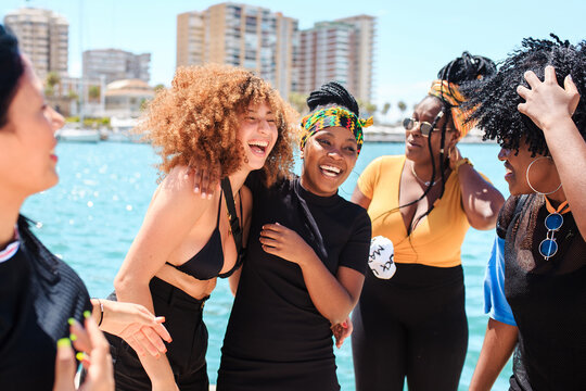 Company of diverse female friends laughing during city stroll near waterside in summer