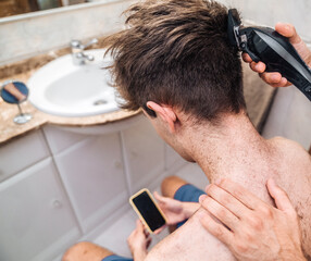 From above back view of crop shirtless male focusing on screen and watching tutorial video after self cutting hair in light bathroom at home as concept bad decisions or result of self isolation