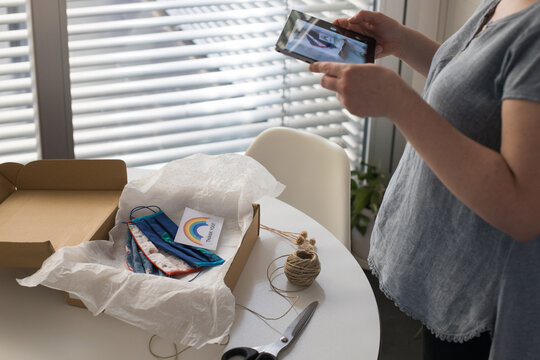 From above of crop faceless female using tablet while standing near round table with cardboard box with collection of handmade masks for COVID 19 pandemic