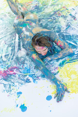 Top view of a naked woman in a spray of paint. A girl without clothes lies on a white floor in color stains.