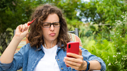 Concentrated thoughtful young woman in headphones using smartphone sit on chair in garden outdoor,writing notes.Focused female student watching online webinar,learning language,listening audio course