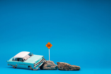 Light blue, retro car figurine next to two boulders and a construction zone sign, on blue...