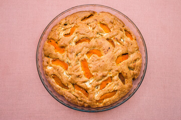 A large, round baked apple and apricot pie stands in a glass form on a table with a tablecloth.