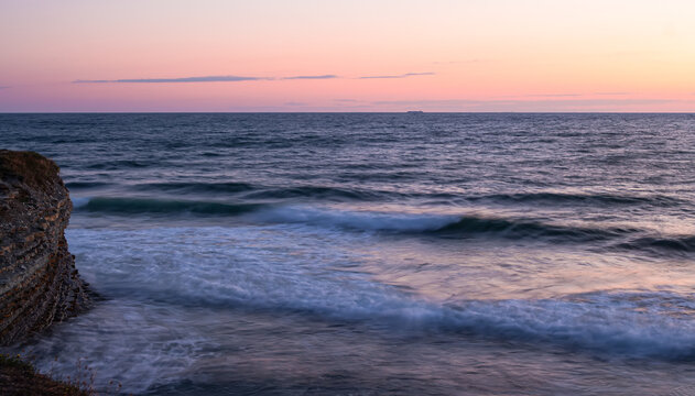 Soft pink sea sunset. Photo at a long shutter speed of one second. Smooth waves beat against the rocks. Cargo ships and tiny clouds can be seen on the horizon.