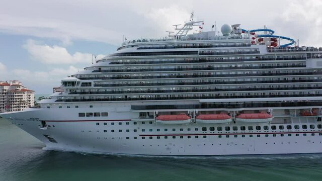Miami, May 2019. Carnival Cruise liner sailing to the Atlantic ocean. 4K aerial view of white cruise ship's board leaving cruise terminal. People on upper deck of the ship. Water transportation