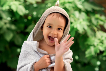 Smiling toddler boy wrapped in big bathing towel after swimming in outdoor pool. Happily laughing child waving hand. Green leaves in background. Summer activities for children concept