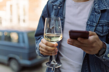 Young man in blue jacket holding glass of white wine and texting message on cell phone. Copy space, summertime relax concept.