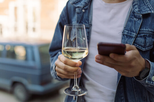 Young man in blue jacket holding glass of white wine and texting message on cell phone. Copy space, summertime relax concept. - Powered by Adobe