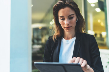 portrait professional female banker standing near office in evening street communicates online by tablet. Businesswoman pushing message to colleagues using computer  on background lights