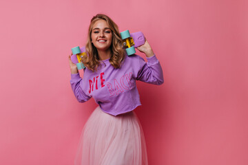 Enchanting female model in white skirt posing with longboard. Indoor photo of glad blonde girl with skateboard isolated on pastel background.