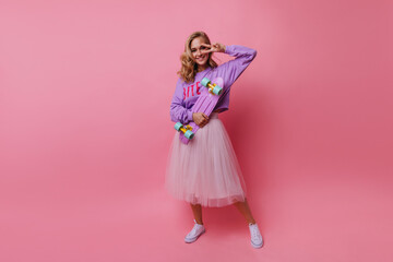 Full-length shot of trendy woman in long white skirt posing with peace sign. Indoor photo of smiling pretty girl with pink skateboard.