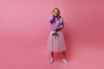 Excited charming girl in long lush skirt posing on pink background. Indoor photo of stylish female model with purple skateboard.