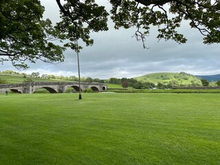 Fototapeta na wymiar Landscape view, across the village green in Burnsall, with the old bridge straddling the river Wharfe in, Burnsall, Skipton, UK