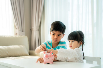 Two Asian cute brother and sister insert coin  in piggy bank together on the table in living room at home. Concept of bonding of sibling, friendship and saving, investment money concept