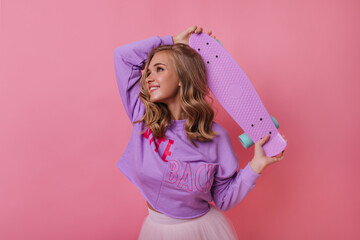 Jocund pretty girl looking up with cute smile. Blonde beautiful woman holding skateboard in studio.