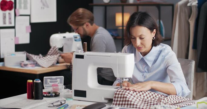 Beautiful woman and bearded man sitting at table and working on sewing machines. Fashion designers tailoring new clothing collection at studio.