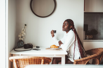 A young authentic black woman with long braids is having breakfast in the morning: sitting on a cane-chair at a cozy table with a cup of coffee and a croissant and pensively looking outside the window