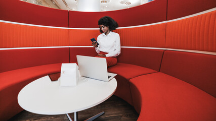 Beautiful African-American woman entrepreneur is sitting in a cozy office coworking area and having business talks via her smartphone and the laptop; a copy space place on the right for your ad