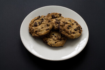 Four chocolate chip cookies on white plate on black background.