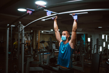 Muscular young fitness man with medical mask doing exercises on horizontal bar in a gym club. COVID 19 coronavirus protection
