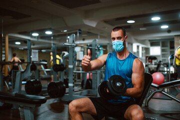 Fototapeta na wymiar A young muscular caucasian athlete with a mask on his face, with a weight in his hand gives a thumb up in the gym. Protection against COVID - 19 coronavirus
