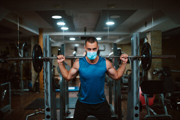 A young caucasian athlete man with a mask on his face exercises and lifts weights in the gym. COVID 19 coronavirus protection