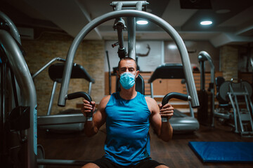 Portrait od young muscular fitness man with medical mask doing heavy weight exercise for body on machine in the gym.Prevention of COVID - 19 coronavirus