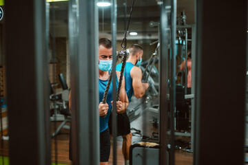 Obraz na płótnie Canvas A handsome young caucasian man with a face mask is exercising in the gym on a device. Protection against COVID - 19 coronavirus