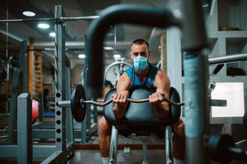 A young caucasian athlete man with a mask on his face exercises and lifts weights in the gym. COVID 19 coronavirus protection
