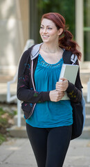 Beautiful female student, with red hair, wearing backpack on campus - walking and holding large textbook