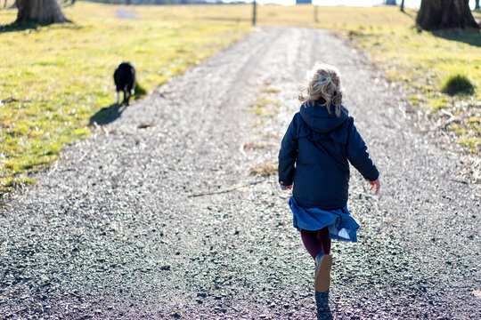 Young Child From Behind Running In The Country With Her Dog Up Her Driveway Towards The Fields
