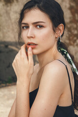 Close-up portrait of a beautiful young woman with curly brown hair tied in ponytail, mysterious eyes looking at you through the lens. Dressed in black top with silver accessories