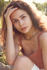 Close-up portrait of a beautiful young woman with curly brown hair, mysterious eyes and big red lips, looking at you through the lens. The sun touches her from behind and creates a pleasant feeling.