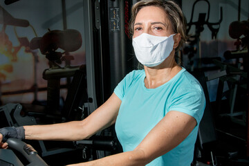 Fit Lady Working out on a Stationary Bike Wearing a Protective Mask 