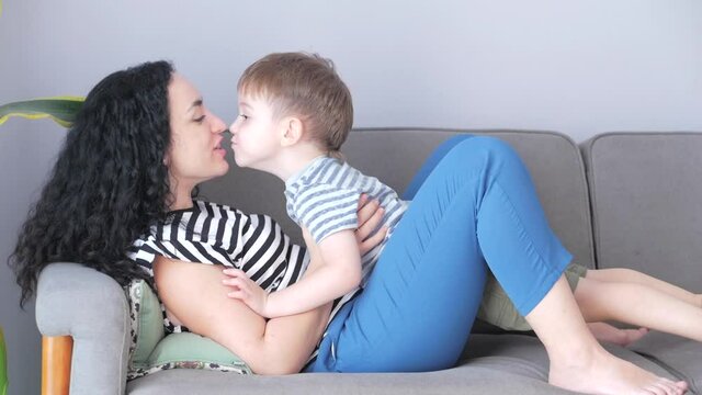 Happy healthy family young mom hugs cute little child son who kisses mother on lips or cheek sitting on sofa at home, funny mother and son kid boy love.