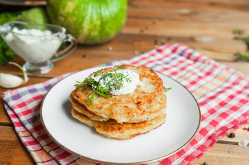 fried zucchini vegetarian pancakes with white sauce and Punch onporcelain plate and red checkered napkin, fresh zucchini and squash, gravy boat on blurred background. simple country food