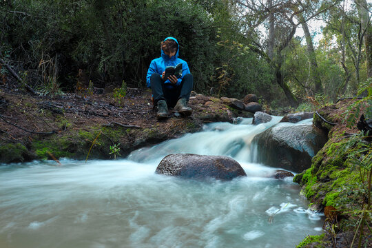 foto de larga exposici&oacute;n de un peque&ntilde;o arrollo con rocas y un tronco en medio en la orilla un hombre joven con cortavientos azul leyendo un libro 